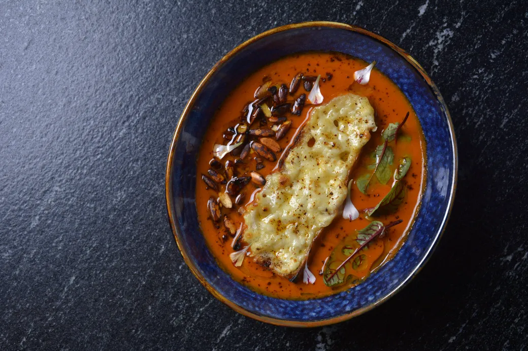 Crema de jitomate ahumado en un plato hondo azul, con crotón gratinado y pepitas